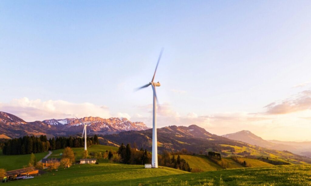 A wind turbine stands tall against a mountainous backdrop under a vibrant sky at sunset.